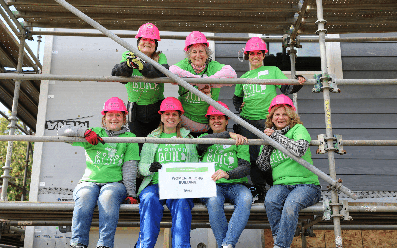 Seven women sitting and standing on scaffolding for a group picture wearing pink hard hats and green Women Build shirts
