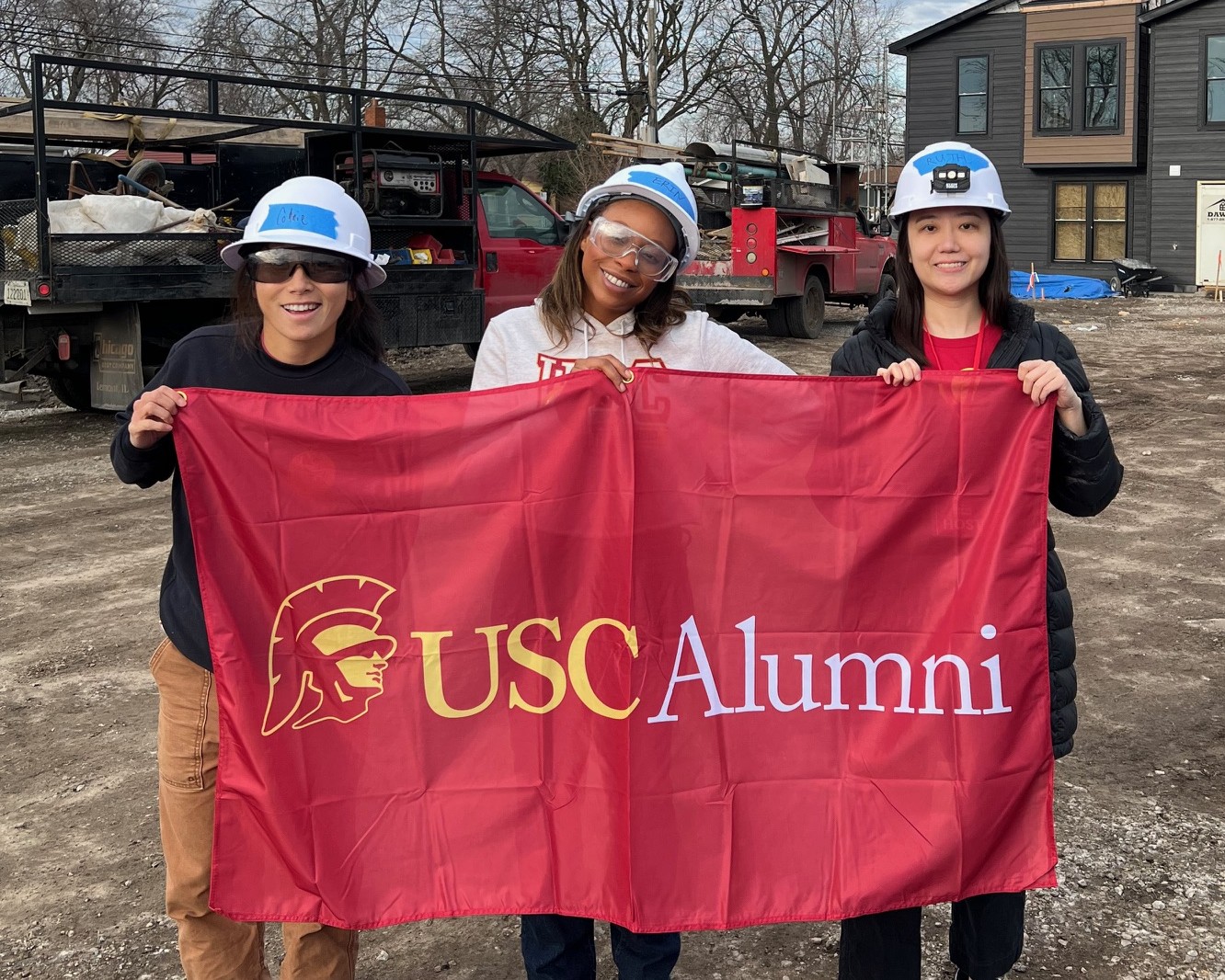 Three people holding a red "USC Alumni" flag on an inactive construction site