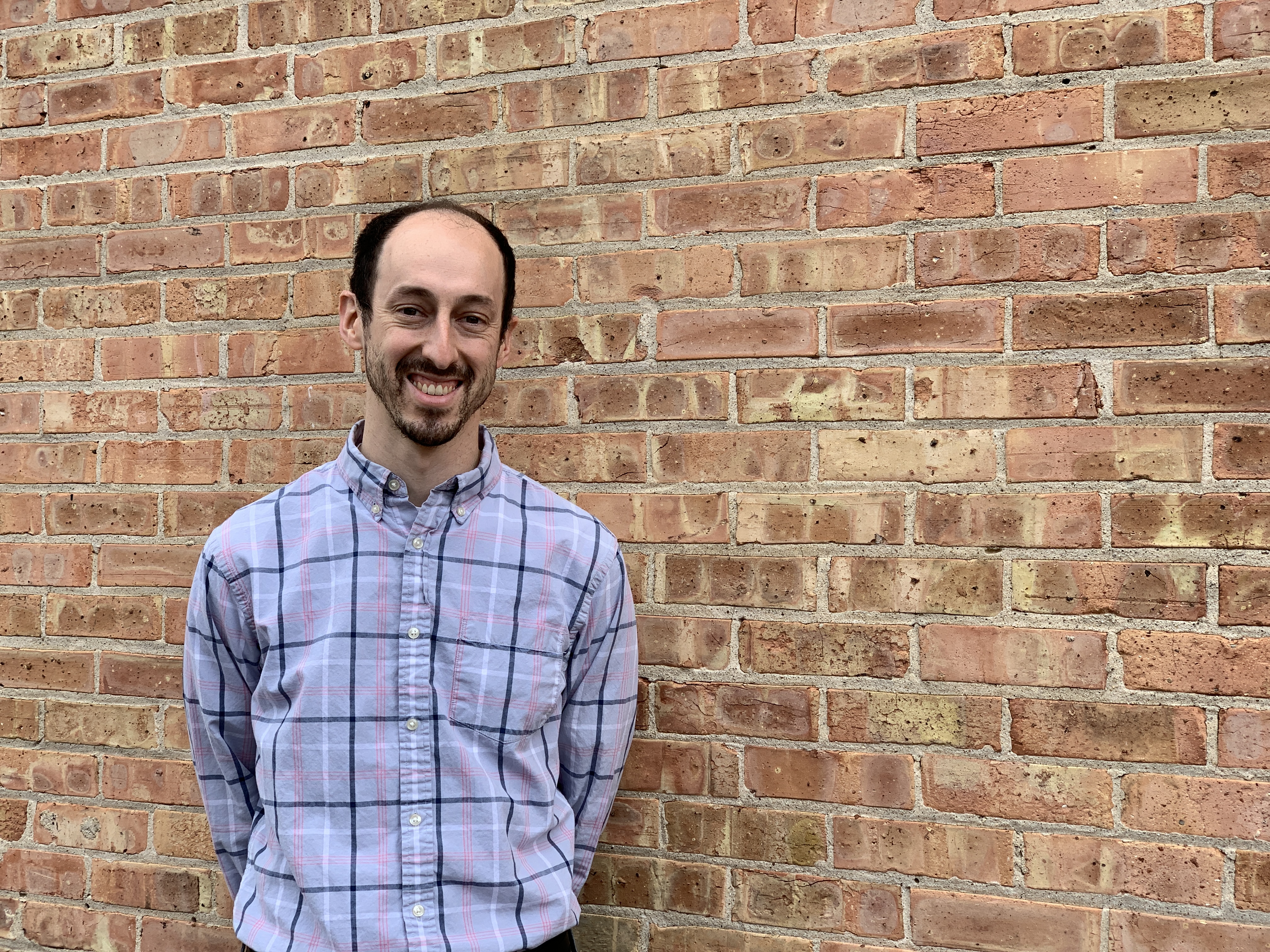 Man in plaid smiling at camera in front of brick wall