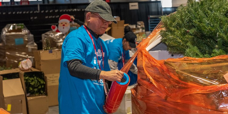 Person with blue shirt and hat wrapping a tall stack of items with orange plastic wrap