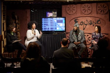 Four people sitting in a panel with a TV in the middle. Woman, second from the left, speaking into the mic, and everyone intently listening.