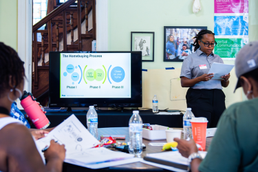 Instructor reading for their notes, standing next to a TV screen that has says, "Homebuying Process"