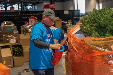 Person with blue shirt and hat wrapping a tall stack of items with orange plastic wrap