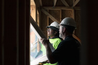 Two people installing a window in an unfinished home; one's face is illuminated by the light outside