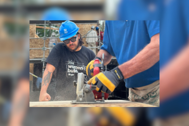Man in black shirt and blue hard hat watching person using a hand saw