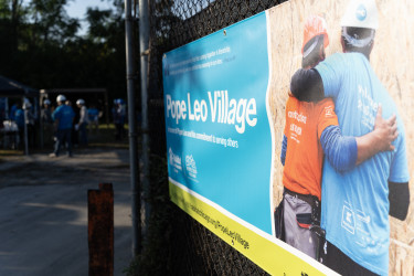 Vinyl sign hung on construction fence that says "Pope Leo Village" with a picture of two people hugging