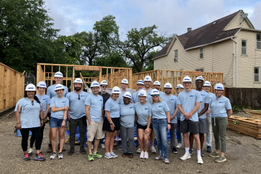 Group in light blue and hard hats on a build site with a framed house behind them