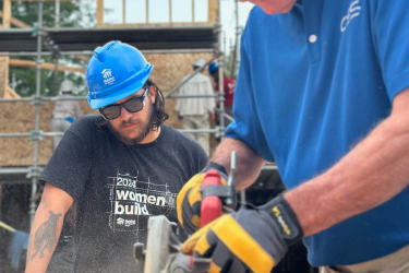 Man in black shirt and blue hard hat watching person using a hand saw