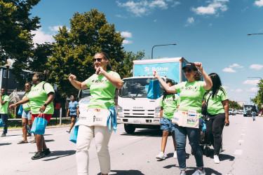A group of Habitat for Humanity Chicago staff in green ReStore shirts dance in front of a blue ReStore truck in the middle of a wide street. Green trees and blue sky can be seen in the background.
