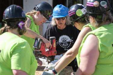 Five women in hard hats and safety glasses are circled around a piece of wood on a stand. One woman is holding a nail gun pressed to the wood, while another woman in a black shirt and blue hard hatappears to be instructing.